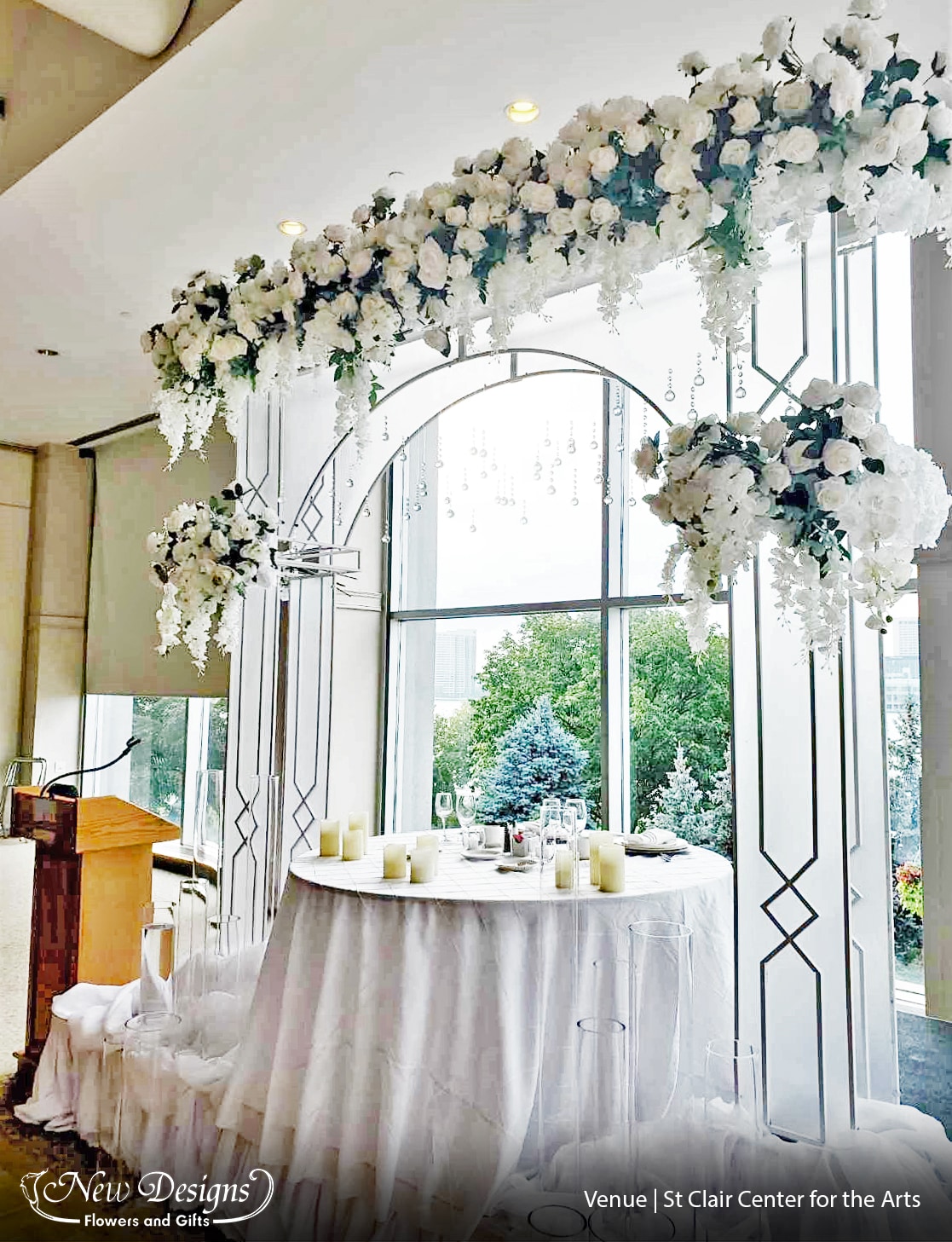 An elegant 8 foot white metal arch, with frosted panels, stands in front of large windows. The top and 3/4 sections over flowing with white roses and orchids, Jewels hang down from the middle over a table overflowing with chiffon material